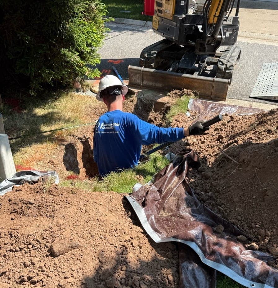 Sweetwater Plumbing technician in an open trench replacing a sewer lateral beside a mini excavator